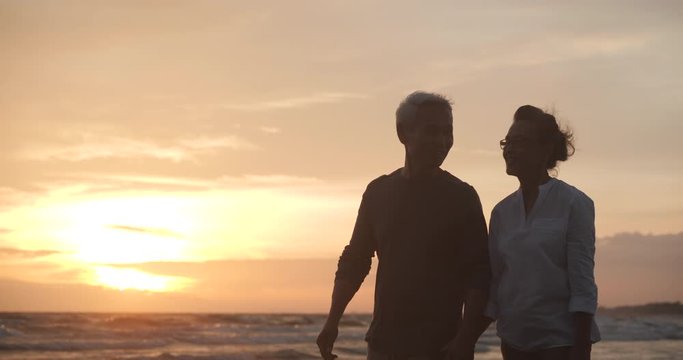 Asian senior couple walking and talking on beautiful tropical beach at sunset in slow motion.