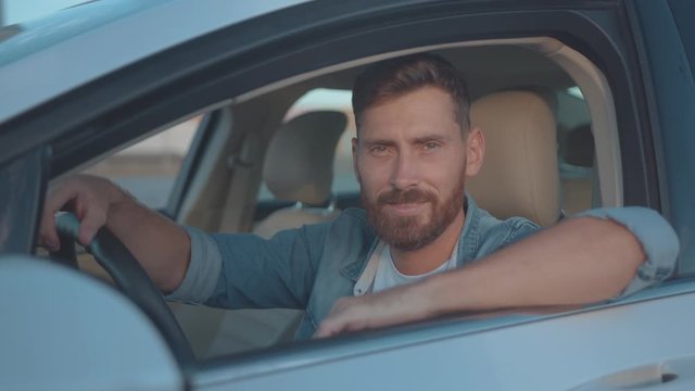 Portrait of nice-looking positive guy sitting in driver seat of modern car transport at sunset and smiling.