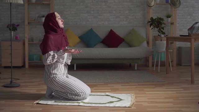 muslim woman in a traditional scarf praying on rug in her living room