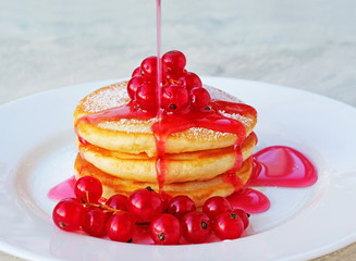 Mini pancakes with red currant and syrup on white plate. Pouring syrup on a stack of pancakes.