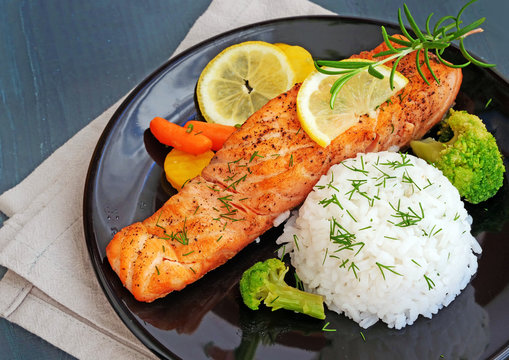 Salmon steak with vegetables, rice and rosemary on black plate over wooden table