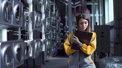 A young pretty white woman works in the paint shop of the plant, she checks the unpainted metall products and making record in check list.