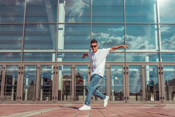 athlete man dancer in white T-shirt, jeans glasses, dancing, summer city, background glass windows of building, active lifestyle, acrobatic stunt break dance, street fashionable artist positive music