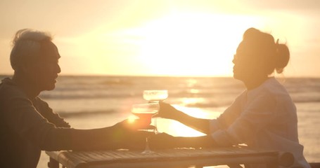 Romantic asian senior couple watching sunset and enjoying a glass of wine on beautiful tropical beach in slow motion.