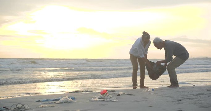 Asian senior couple cleaning up the beach with plastic bags full of garbage. Slow Motion. Safe ecology concept. 4k resolution.