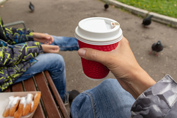 Unrecognizable woman sitting on a bench in park with her family and holading a red paper takeaway cup of hot coffee in her hand, autumn warming drinks and street food concept