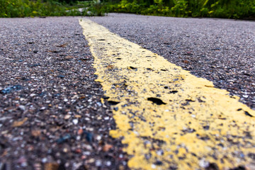 Yellow solid line, grunge single road marking on asphalt close up with rural landscape on background