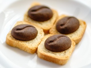 Four brown cookies with shape of cocoa beans lay on square breadcrumbs in white plate.