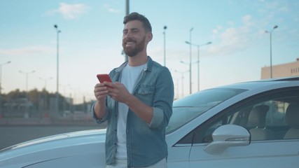 Handsome joyful man using smartphone leaning on car outdoor. Happy smiling male driver messaging on mobile phone looking around staying in modern city. Summertime. - Powered by Adobe