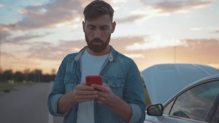 Nervous young man using smartphone calling car assistance services standing by breakdown automobile on the roadside at sunset.