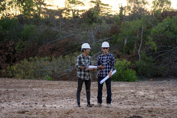 portrait of two engineer's or architect's with hardhat walking across the field with blueprint tablet computer and plant for build something