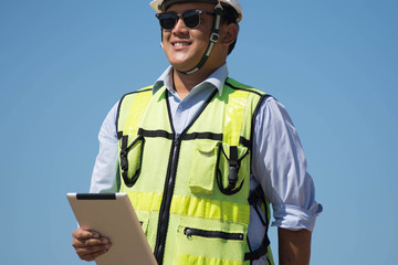 bottom rear view of male engineer walking and holding tablet computer on bright blue sky for planing a construction dress with protective vest, hard helmet and