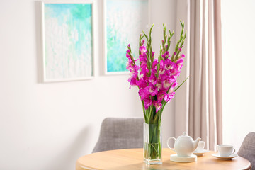 Vase with beautiful pink gladiolus flowers and tea set on wooden table in room, space for text