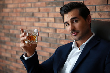 Young man with glass of whiskey near brick wall indoors
