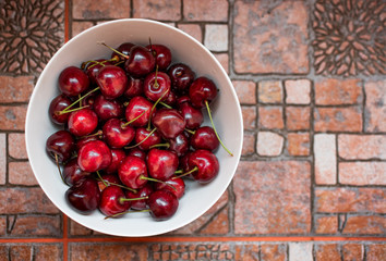 Top view of a white bowl full of ripe cherries covered with drops of water, over blurred background of kitchen tiles.