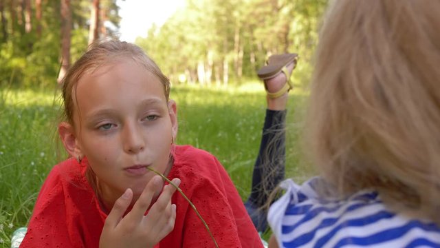 Happy childhood and outdoor activities. Two playful teen girls lies on grass and having a fun in nature park together. Girl is eating grass and talking with her friend. Children friendship.