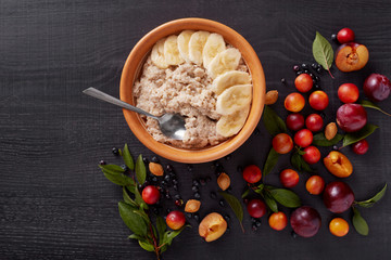 Image of oatmeal with banana, alich, pluns, bluberries, pits and green leaves on dark wooden surface. Delicious and healthy breakfast, table with vitamins, careal with fruit. Healthy eating concept.