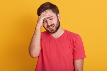 Tired young man keeps hand on forehead, handsome guy wearing red casual t shirt, posing with closed eyes over yellow studio background, suffers from headache. Stress or problem concept. Front view.