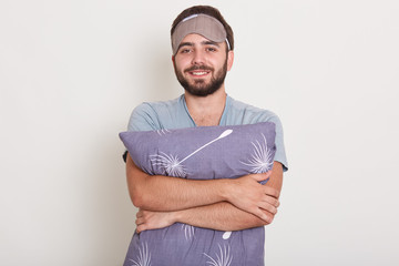 Close up portrait of man dressed gray casual t shirt ready to go to bed, white wall on background. Guy with happy face holds pillow. Macho with beard and mustache relaxing, having nap, resting at home