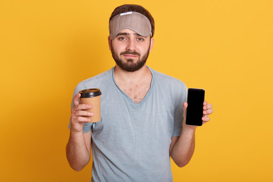 Portrait Of Upset Handsome Bearded Young Hipster Man In Gray T Shirt And Blindfold Standing With Coffee In One Hand And Phone With Blank Screen In Another, Isolated Over Pink Studio Background.