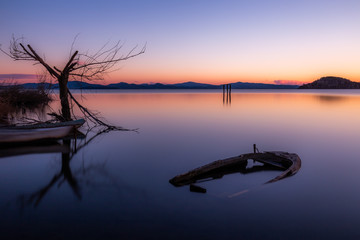 An almost completely sinked little boat in Trasimeno lake (Umbria, Italy) at dusk, near a skeletal tree