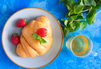 Tasty croissant with  fresh strawberries and green tea on blue cement background.Top view.