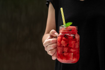 Girl holds in hand summer watermelon drink in a glass with slices of watermelon on a dark background
