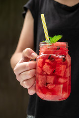 Girl holds in hand summer watermelon drink in a glass with slices of watermelon on a dark background