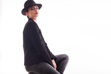 Young fashion model with a stylish hat and jacket posing sitting on a char in studio over white background