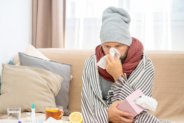 Obraz na płótnie Canvas Sick man wearing grey hat and scarf, blowing nose and sneeze into tissue. Male have flu, virus or allergy respiratory. Healthy, medicine and people concept. Headache and fever remedies.