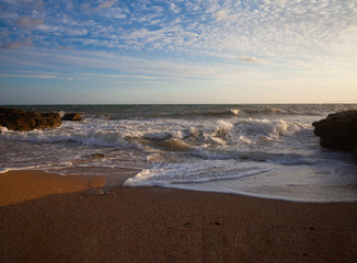 Beautiful сloudy Blue dawn over sea and waves hitting and splashing rocky stone beach with sand
