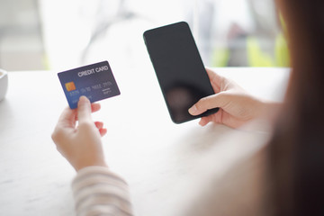 Closed up and focus on hand of young woman hold a credit card and other hand using a smart phone which is shopping online and sitting on table near big mirror at cafe. online payment concept.