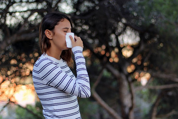 Health and medicine concept - Young woman blowing nose into tissue on the forest.