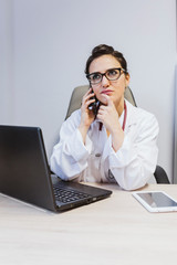 young doctor woman working on laptop at the consult. Talking on mobile phone and looking worried. Modern Medical concept indoors