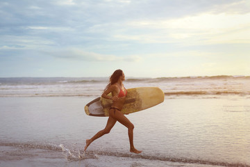 A young woman with white surfing in her hands running along the ocean shore at sunset.