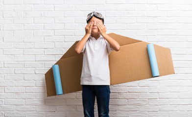 Boy playing with cardboard airplane wings on his back covering eyes by hands