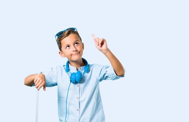 Kid with sunglasses and headphones traveling with his suitcase standing and thinking an idea on blue background