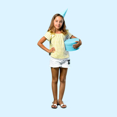 Full body of Little girl at a birthday party holding a gift posing with arms at hip on blue background