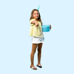 Full body of Little girl at a birthday party holding a gift points finger at you on blue background