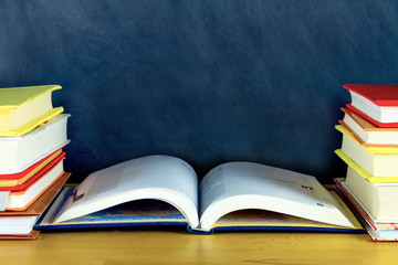 Stacks of colorful books are on either side of desk. Open notebook is on center of table in classroom. Chalkboard blackboard on background. Back to school, university, college concept. Autumn mood.