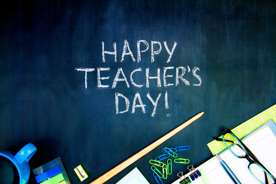 Back to school, college, university concept. Congratulatory inscription of happy teacher's day on blackboard background from pupils. Office supplies, pointer, glasses, notebook are scattered on canvas
