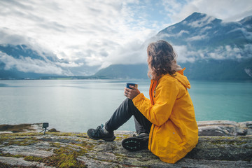 A young beautiful girl is drinking tea on the shore of the fjord, a trip to Norway, the Nordic...