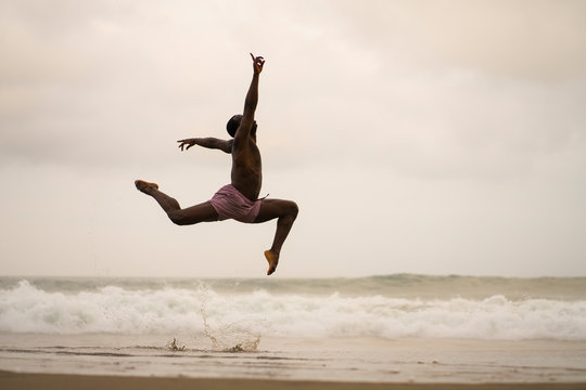 dramatic contemporary dance choreographer doing ballet beach workout . young attractive and athletic afro black American man dancing on sunrise jumping