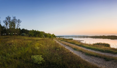 Rural panoramic landscape as a country road separates the lake from the forest. Beautiful evening scene, calm autumn background near a meadow of steppe vegetation.