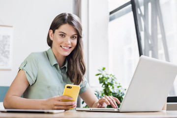 Woman using laptop and smart phone in office. Beautiful girl at desk looking at camera. Entrepreneur, businesswoman, freelance worker, student working on computer. Business, technology concept