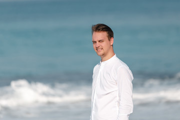 Outdoor summer portrait of smiling happy man in  white shirt on the beach near the sea looking at the camera. Groom on the beach