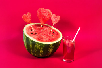 Useful watermelon juice with a tube and a large ripe watermelon decorated with hearts on sticks on a red background. Concept of summer mood, harvest of seasonal berries and fruits