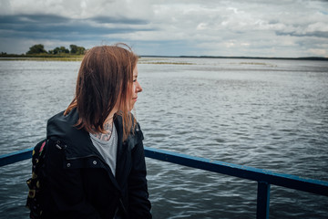 Attractive girl looking to the side standing near the water. Woman looks thoughtfully into the distance, standing on the street on a cloudy day