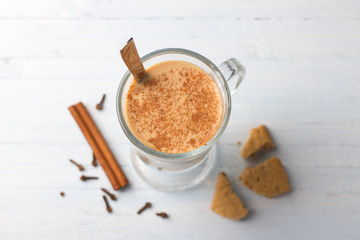 Spicy warming tea with milk in a glass cup and cinnamon sticks, cloves, with gingerbread cookie on light gray blue background