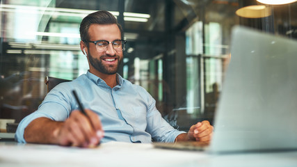 Positive talk. Young and cheerful bearded man in eyeglasses and headphones talking with client by phone and smiling while sitting in the modern office